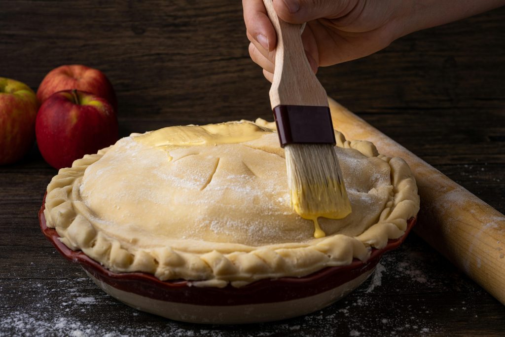 A close-up view of a homemade apple pie being prepared with an egg wash on a wooden surface.